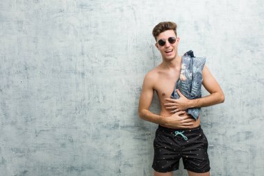 Mayo giyen genç beyaz adam mutlu bir şekilde gülüyor ve ellerini karnının üstünde tutarak eğleniyor..