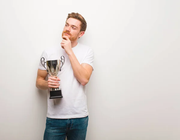 Young redhead man doubting and confused. Holding a trophy.