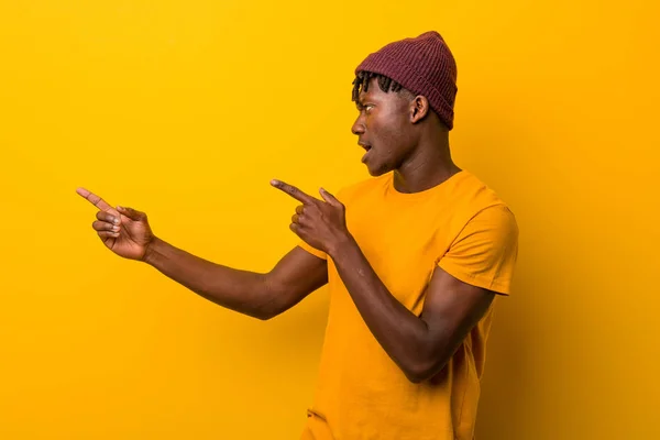 Young black man wearing rastas over yellow background excited pointing with forefingers away.
