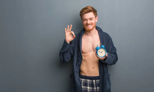 Young redhead man wearing pajama cheerful and confident doing ok gesture. He is holding an alarm clock.