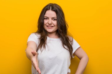 Genç, kıvrımlı, iri yarı bir kadın selam vermek için kameraya elini uzatıyor..