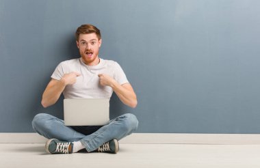 Yerde oturan genç kızıl saçlı öğrenci şaşırdı, başarılı ve başarılı hissediyor. Elinde dizüstü bilgisayar var..