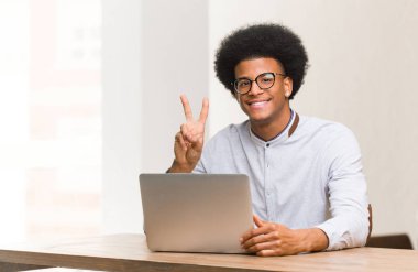 Genç siyah adam dizüstü bilgisayarını eğlenceli ve mutlu bir şekilde kullanarak zafer jesti yapıyor.