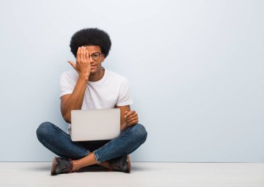 Genç siyah bir adam dizüstü bilgisayarla yerde oturmuş mutlu bir şekilde bağırıyor ve yüzünü eliyle örtüyor.