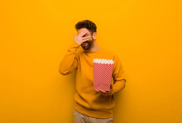 Young man holding popcorns embarrassed and laughing at the same time