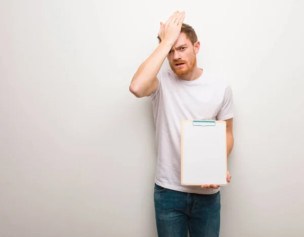 Young redhead man worried and overwhelmed. He is holding a clipboard.