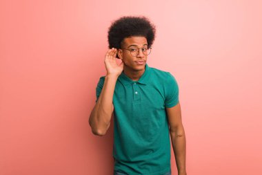 Genç Afro-Amerikan bir adam pembe bir duvarın ardında bir dedikoduyu dinlemeye çalışıyor.