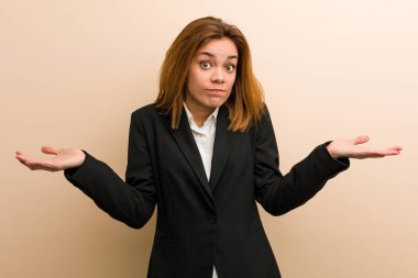 Young caucasian business woman doubting and shrugging her shoulders in questioning gesture.