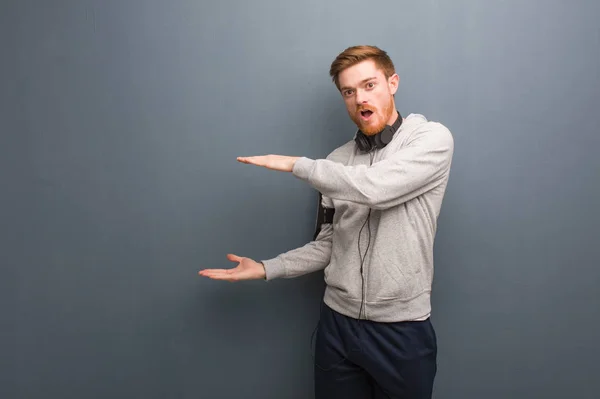Young fitness redhead man holding something very surprised and shocked