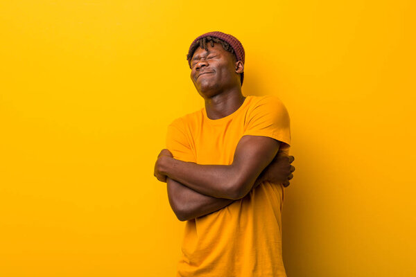 Young black man wearing rastas over yellow background hugs himself, smiling carefree and happy.