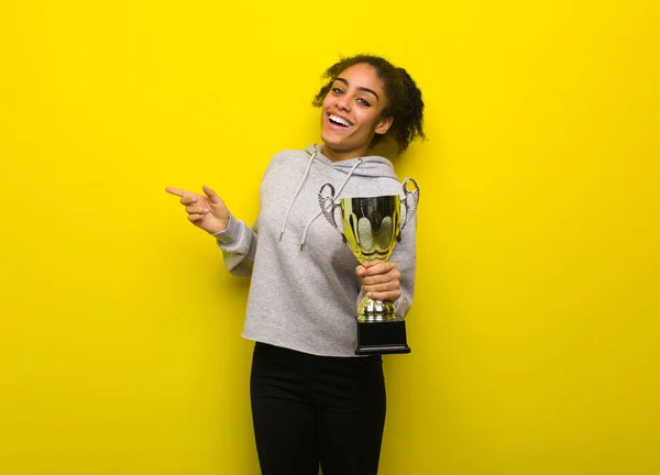 Young fitness black woman pointing to the side with finger. Holding a trophy.