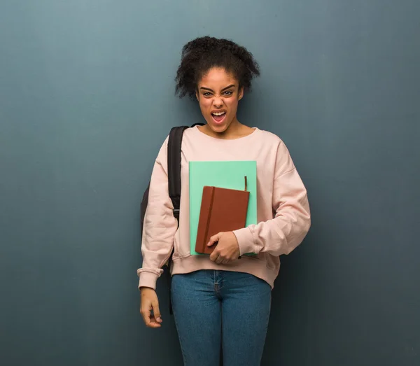 Young student black woman screaming very angry and aggressive. She is holding books.