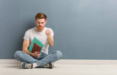 Genç, kızıl saçlı bir öğrenci yerde oturuyor ve kızgın bir ifadeyle yumruğunu gösteriyordu. Elinde kitaplar var..