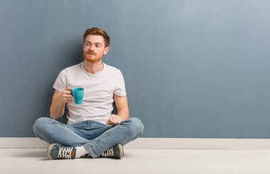 Genç kızıl saçlı bir öğrenci yerde oturmuş kendine güvenen ve kollarını kavuşturup yukarı bakıyor. Elinde bir kahve fincanı var..