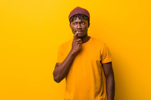 Young black man wearing rastas over yellow background looking sideways with doubtful and skeptical expression.