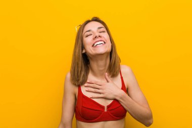 Young caucasian woman wearing bikini and sunglasses laughs out loudly keeping hand on chest.