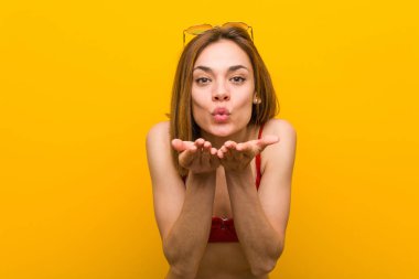 Bikini ve güneş gözlüğü takan, dudak büken ve hava öpücüğü vermek için avuç içi tutan beyaz bir kadın..