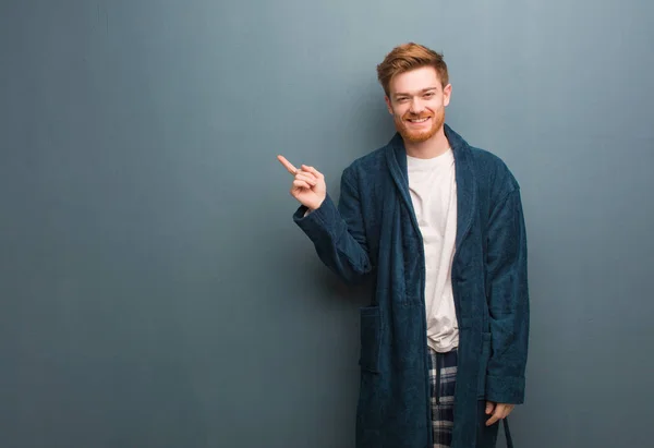 Young redhead man in pajama pointing to the side with finger