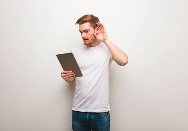 Young redhead man try to listening a gossip. Holding a tablet.