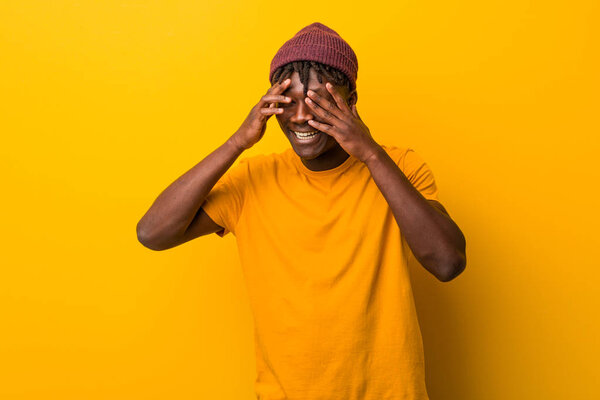 Young black man wearing rastas over yellow background blink through fingers frightened and nervous.