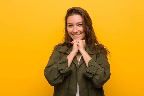 Young european woman isolated over yellow background keeps hands under chin, is looking happily aside.