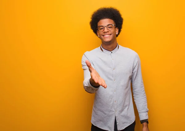 Young african american man over an orange wall reaching out to greet someone