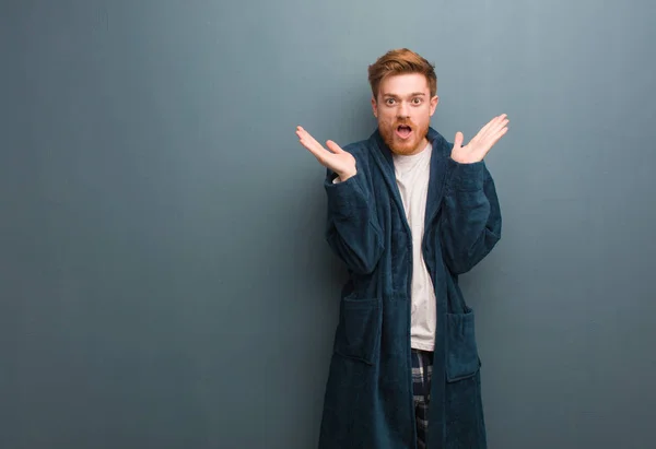 Young redhead man in pajama surprised and shocked