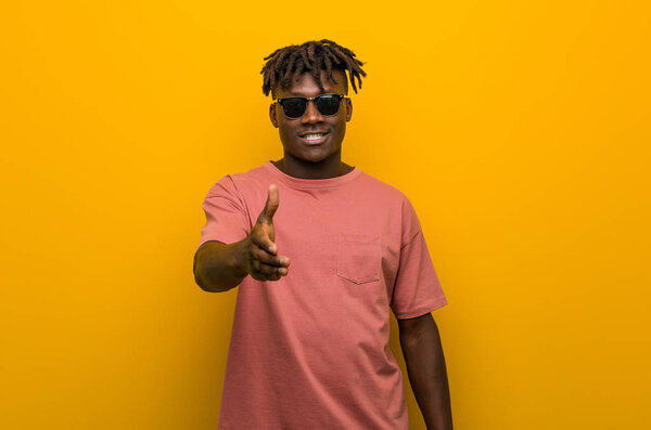 Young casual black man wearing sunglasses stretching hand at camera in greeting gesture.