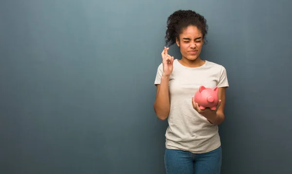 Young black woman crossing fingers for having luck. She is holding a piggy bank.