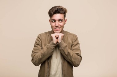 Kahverengi ceket giyen genç beyaz adam ellerini çenesinin altında tutuyor ve mutlu bir şekilde yan tarafa bakıyor..