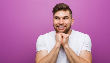 Genç yakışıklı beyaz adam ellerini çenesinin altında tutuyor, mutlu bir şekilde yan tarafa bakıyor..