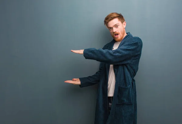 Young redhead man in pajama holding something very surprised and shocked