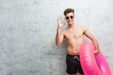 Elinde pembe şişme bir çörek tutan, mayo giyen neşeli ve kendinden emin genç bir adam iyi bir jest yapıyor..