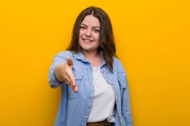 Genç, kıvrımlı, iri yarı bir kadın selam vermek için kameraya elini uzatıyor..