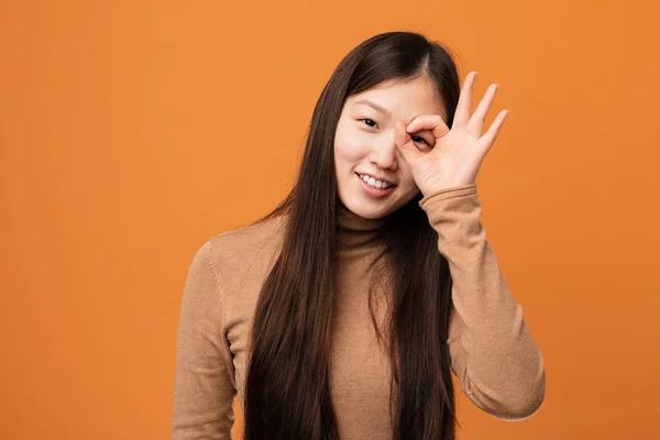 Young pretty chinese woman excited keeping ok gesture on eye.