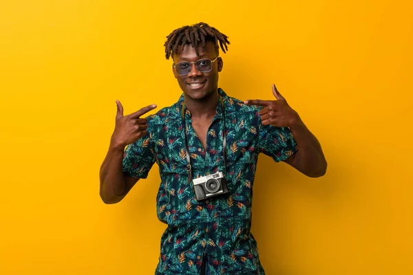 Young black rasta man wearing a vacation look smiles, pointing fingers at mouth.