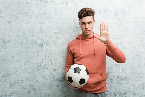 Young sporty man holding a soccer ball standing with outstretched hand showing stop sign, preventing you.
