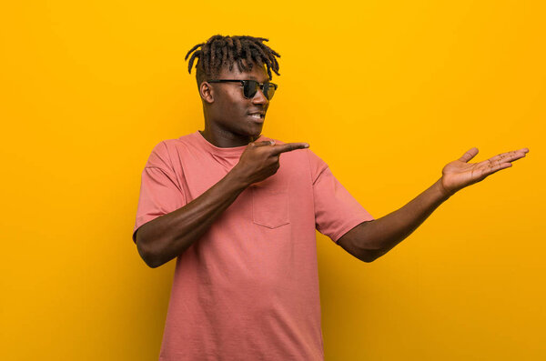 Young casual black man wearing sunglasses excited holding a copy space on palm.