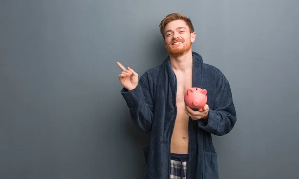 Young redhead man wearing pajama pointing to the side with finger. He is holding a piggy bank.