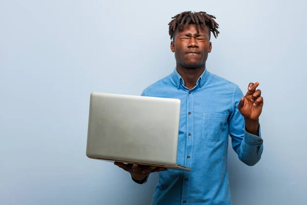 Young rasta black man holding a laptop crossing fingers for having luck