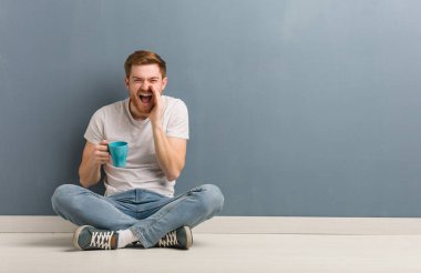 Genç kızıl saçlı bir öğrenci yerde oturmuş, ön tarafa mutlu bir şey bağırıyor. Elinde bir kahve fincanı var..