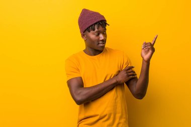 Sarı arka planda rastalar takan genç siyah adam işaret parmağıyla işaret ederek gülümsüyor..