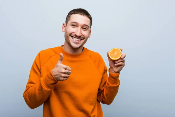 Joven Caucásico Sosteniendo Una Naranja Sonriente Levantando Pulgar Hacia Arriba —  Fotos de Stock