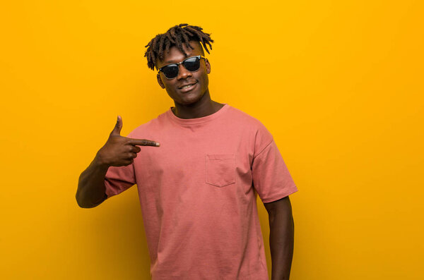 Young casual black man wearing sunglasses person pointing by hand to a shirt copy space, proud and confident