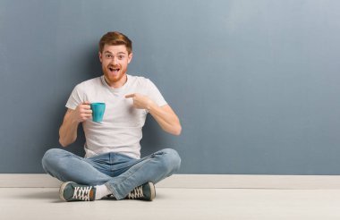 Yerde oturan genç kızıl saçlı öğrenci şaşırdı, başarılı ve başarılı hissediyor. Elinde bir kahve fincanı var..