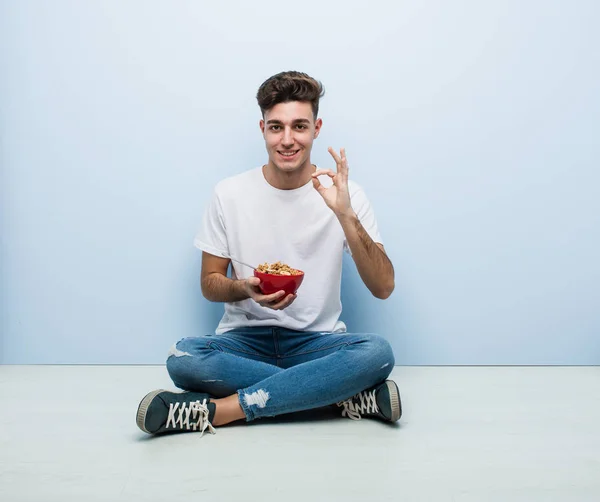 Young man eating cereals sitting on the floor cheerful and confident showing ok gesture.