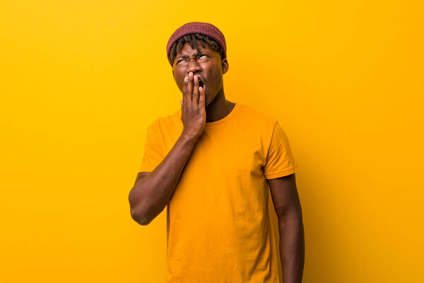 Young black man wearing rastas over yellow background yawning showing a tired gesture covering mouth with him hand.