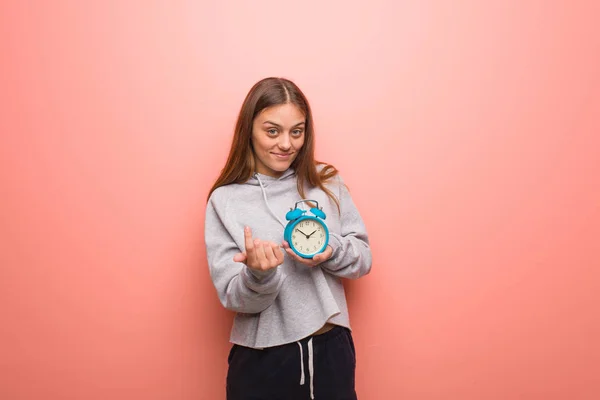 Young pretty caucasian woman inviting to come. She is holding an alarm clock.