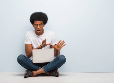 Genç siyahi bir adam dizüstü bilgisayarla yerde oturuyor. İğrenme hareketi yapıyor.