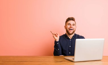 Dizüstü bilgisayarıyla çalışan genç adam işaret parmağıyla işaret ederek gülümsüyor..